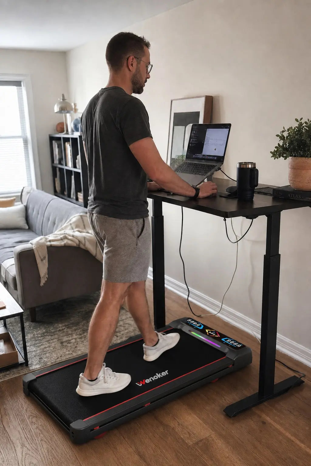 A man working on a laptop at a standing desk while walking on a Wenoker under-desk walking pad treadmill in a home office or living room.
